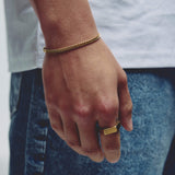 A close-up of a person's hand wearing a gold chain bracelet on the wrist and a gold signet ring on the index finger, paired with a white t-shirt and blue jeans.