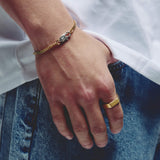 A close-up lifestyle shot of a person's hand resting on blue denim jeans, wearing a gold chain bracelet with a silver lobster clasp and a gold ring.