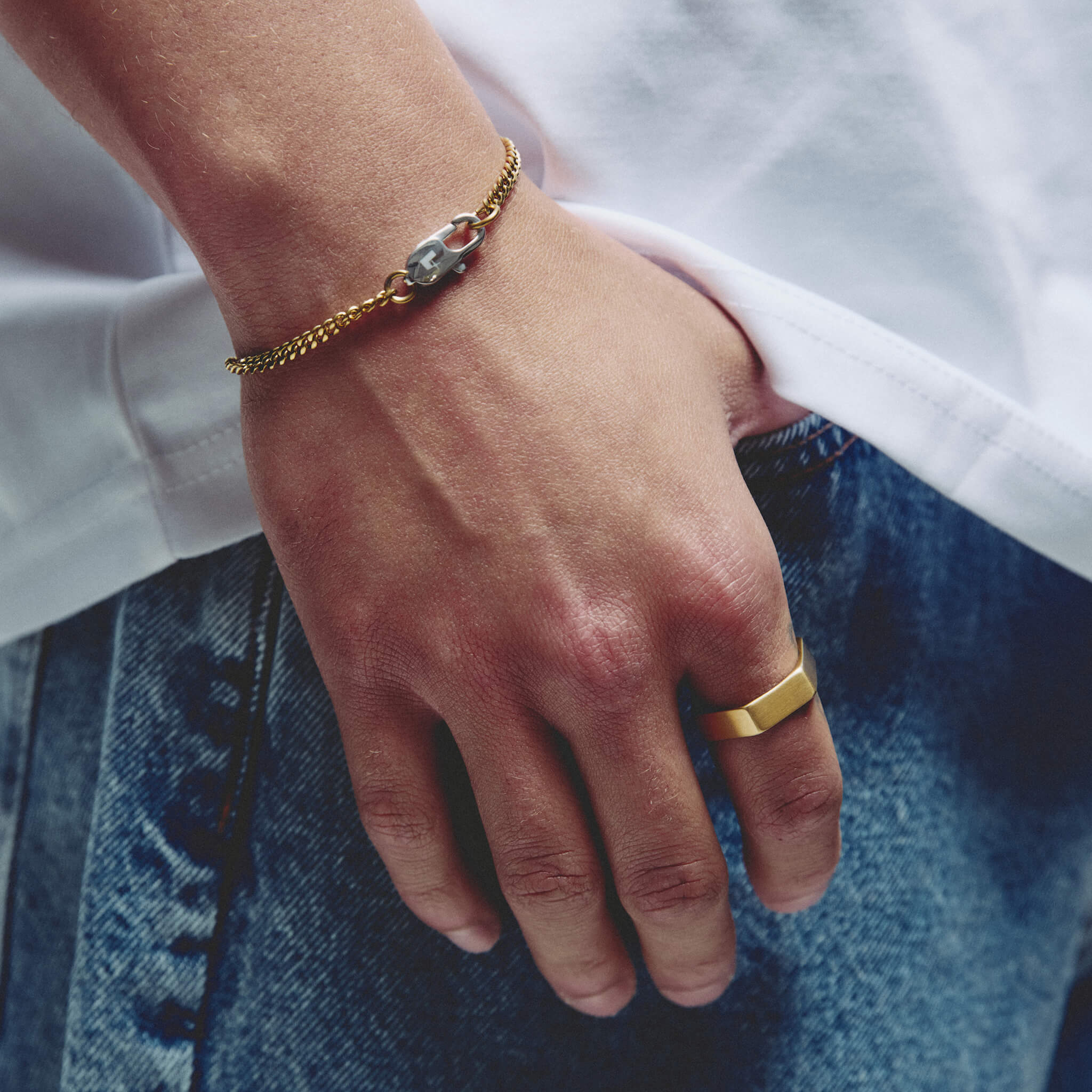 A close-up lifestyle shot of a person's hand resting on blue denim jeans, wearing a gold chain bracelet with a silver lobster clasp and a gold ring.