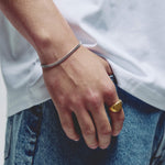 A close-up of a person's hand against their side wearing a silver chain bracelet on the wrist and a gold signet ring on the index finger, dressed in a white t-shirt and blue jeans.