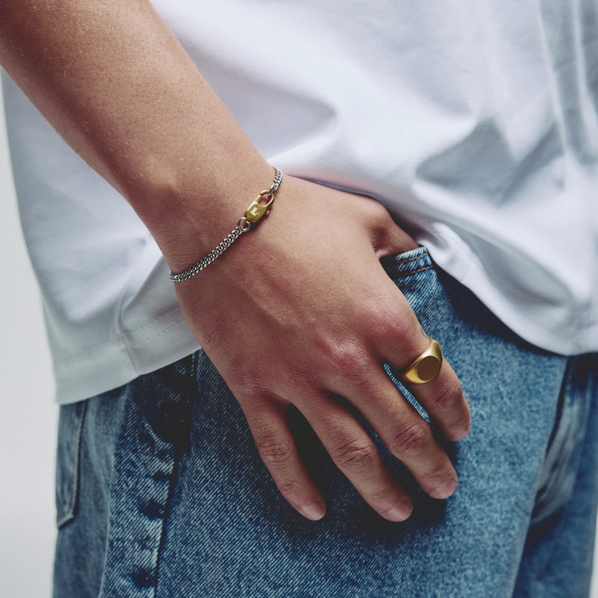 A close-up lifestyle shot of a person's hand resting in their jeans pocket, wearing a silver chain bracelet with a gold lobster clasp charm and a gold signet ring.