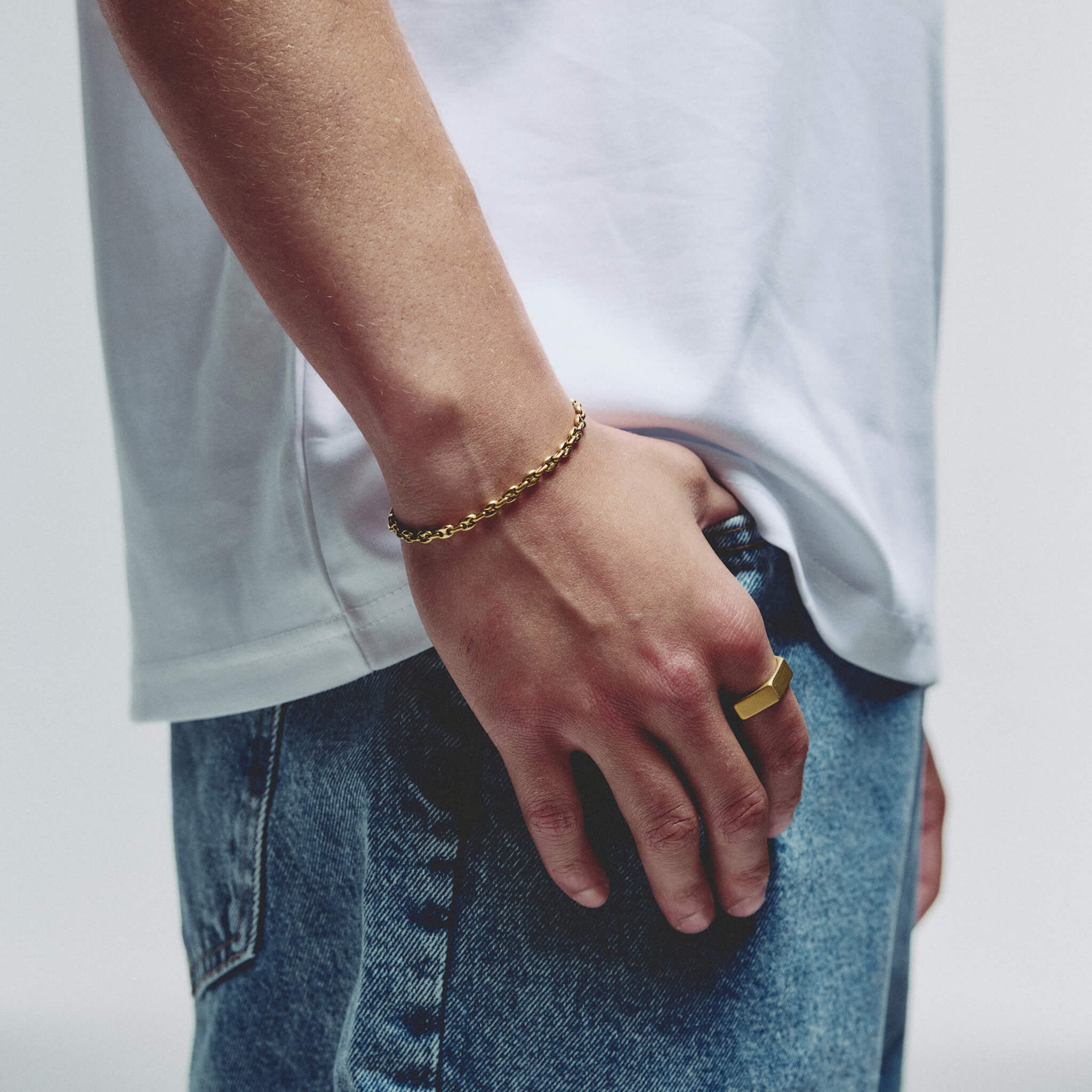 A close-up lifestyle shot of a person wearing a gold chain bracelet on their wrist and a gold ring on their finger, dressed in a white t-shirt and blue jeans against a plain light gray background.
