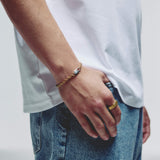 A close-up of a person's hand resting on their thigh, wearing a delicate gold chain bracelet with a silver lobster clasp along with a gold ring on the index finger and blue jeans.