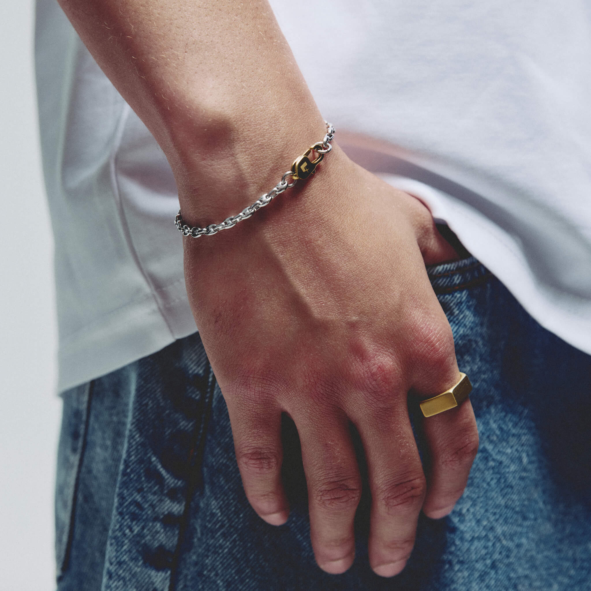 A close-up of a person's hand resting on their jeans, showcasing a silver chain bracelet with a gold lobster clasp on their wrist, along with a gold ring on the finger.