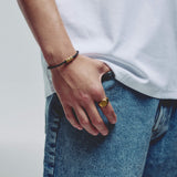 A close-up lifestyle shot of a person's hand resting in their jeans pocket, showcasing a gold signet ring on one finger and a black cord bracelet with a gold hex block magnetic clasp on the other wrist.