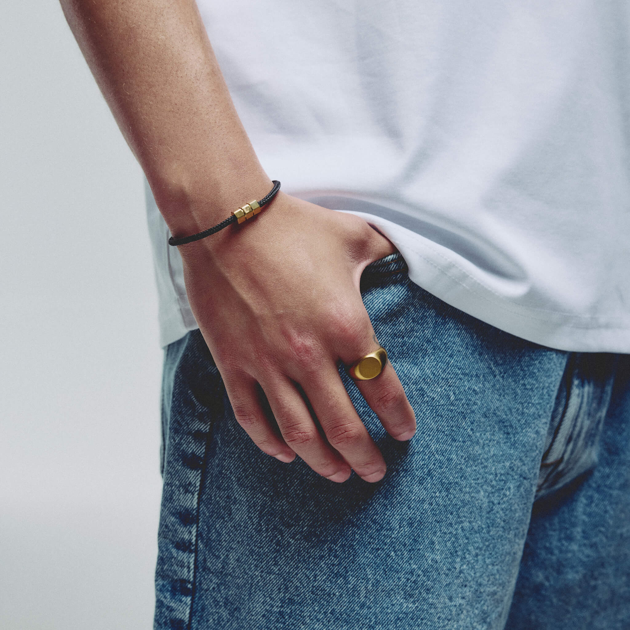 A close-up lifestyle shot of a person's hand resting in their jeans pocket, showcasing a gold signet ring on one finger and a black cord bracelet with a gold hex block magnetic clasp on the other wrist.