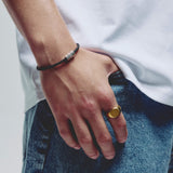 A close-up of a person's hand resting on blue jeans, wearing a black cord bracelet with silver hex block magnetic clap on one wrist and a gold signet ring on the other hand.