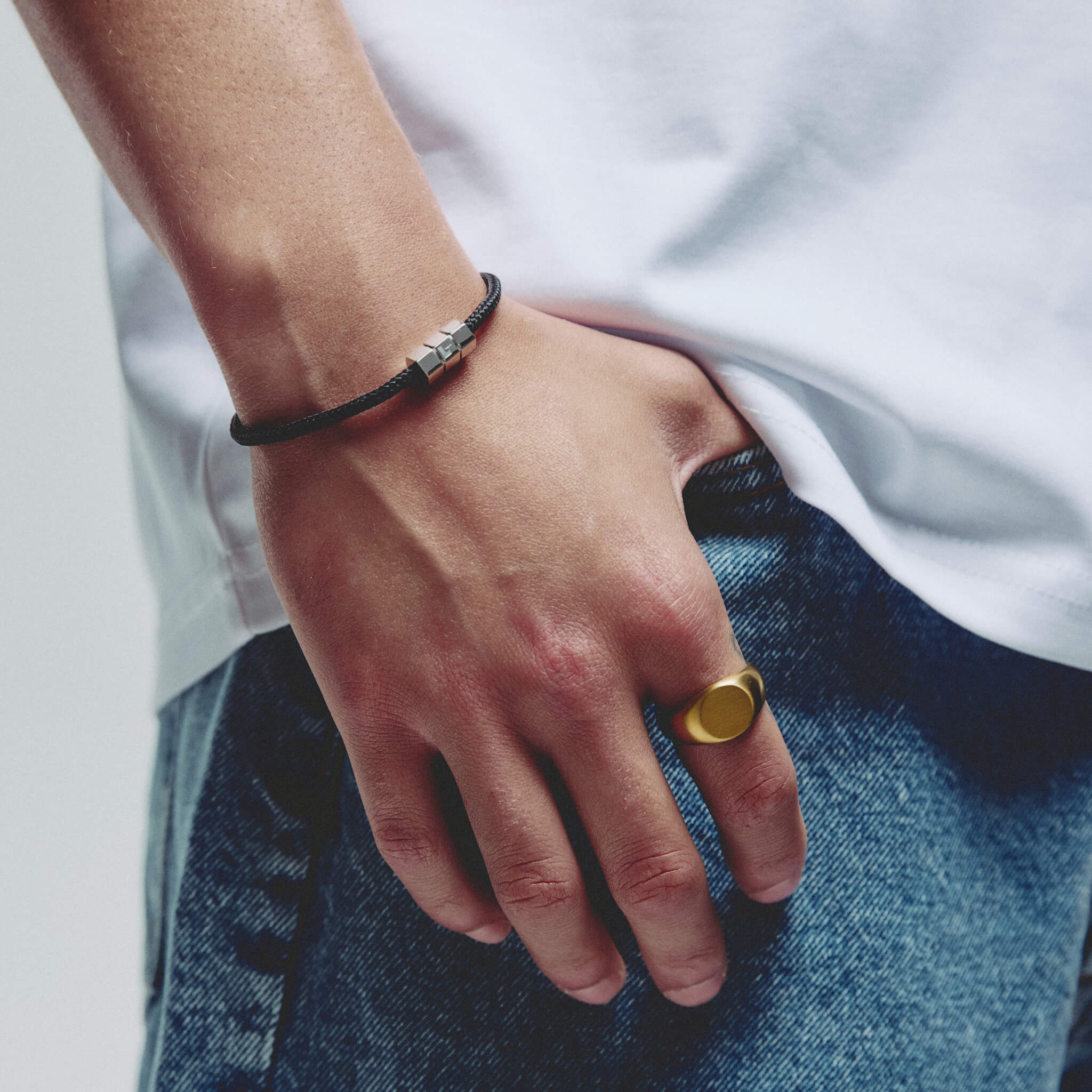 A close-up of a person's hand resting on blue jeans, wearing a black cord bracelet with silver hex block magnetic clap on one wrist and a gold signet ring on the other hand.