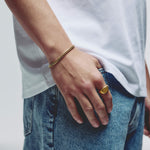 A close-up lifestyle shot of a person wearing a gold chain bracelet on their wrist and a gold signet ring on their finger, styled with a white t-shirt and blue jeans.