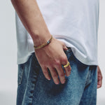 A close-up of a person's hand wearing a gold chain bracelet with a silver lobster clasp paired with a gold ring on the finger, wearing a white t-shirt and blue jeans.