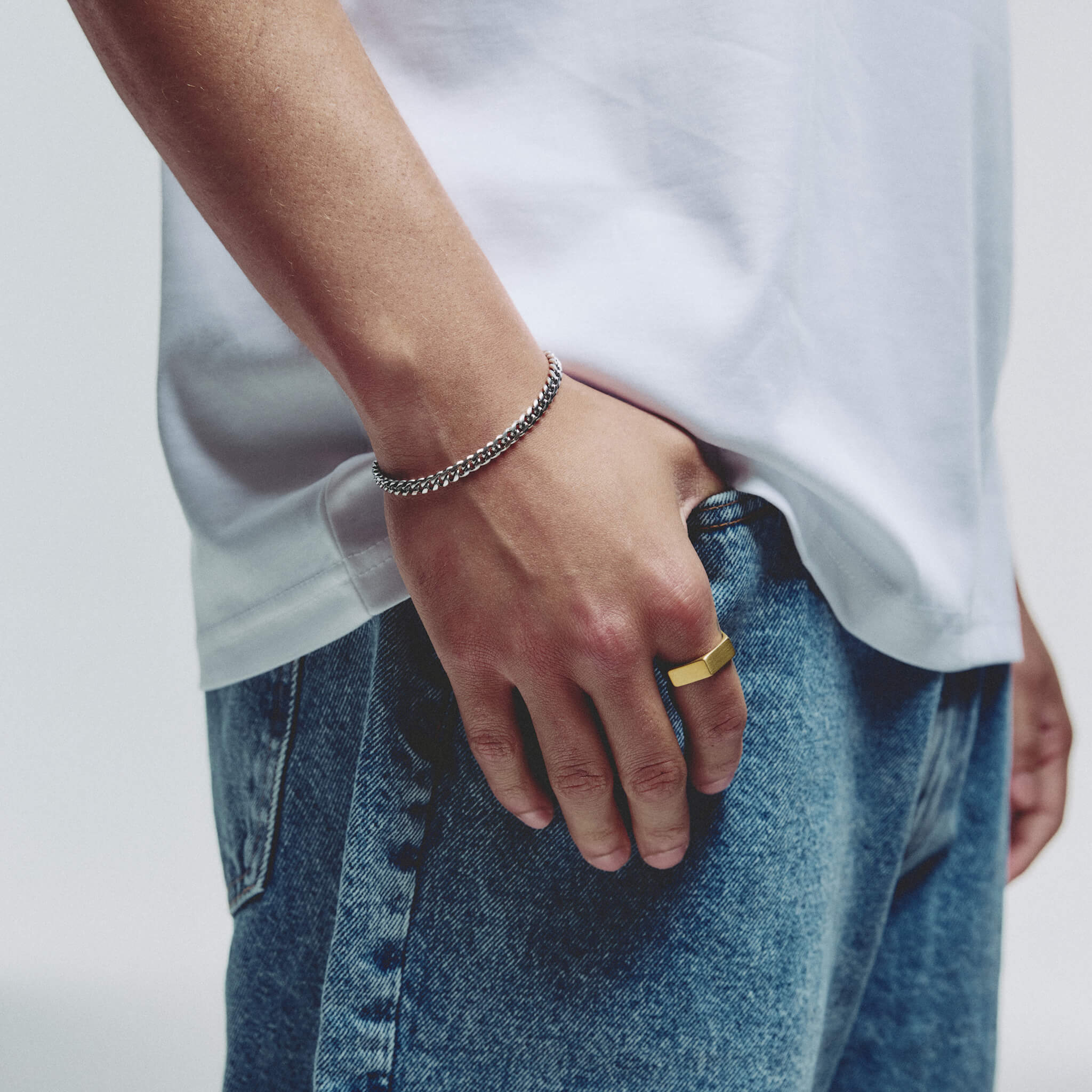 A close-up lifestyle shot of a person wearing a silver chain bracelet on one wrist and a gold signet ring on the other hand, styled with a white t-shirt and blue jeans.
