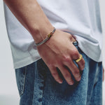 A close-up of a person's hand resting on their thigh, wearing a silver chain bracelet with a small gold lobster clasp and a gold ring.