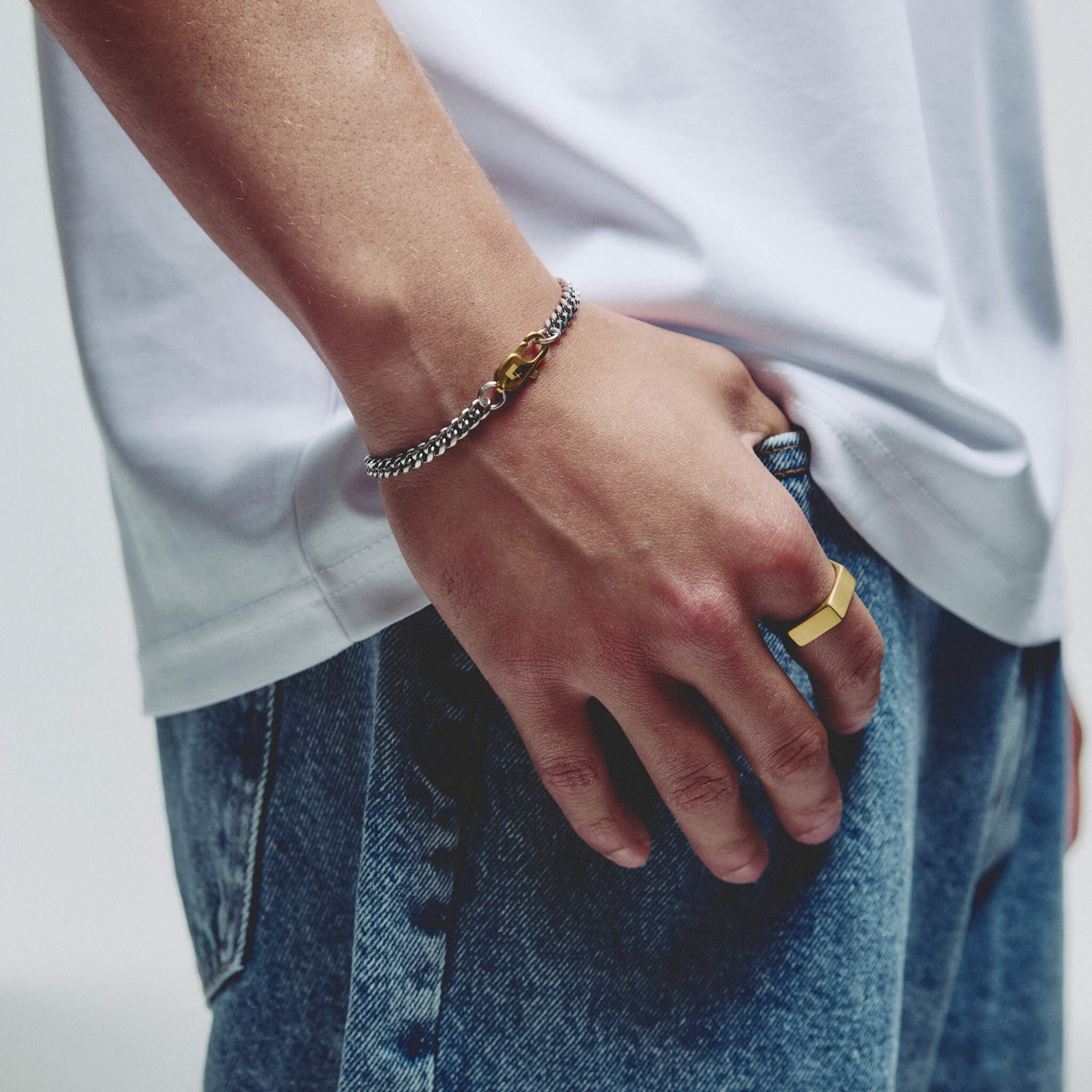 A close-up of a person's hand resting on their thigh, wearing a silver chain bracelet with a small gold lobster clasp and a gold ring.