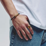 A close-up of a person's hand wearing a gold signet ring on one finger and a black cord bracelet with gold hex block clasp on the wrist, styled with a white t-shirt and blue jeans.