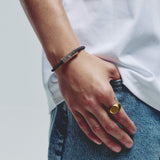 A close-up of a person's hand resting on their hip, showcasing a black cord bracelet with a silver metal hexagonal block clasp that is magnetic, on the wrist and a gold-toned signet ring on the ring finger.
