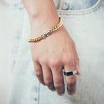 Close-up of hand and wrist featuring thick gold chain bracelet with silver clasp and silver hex style ring on textured fabric background
