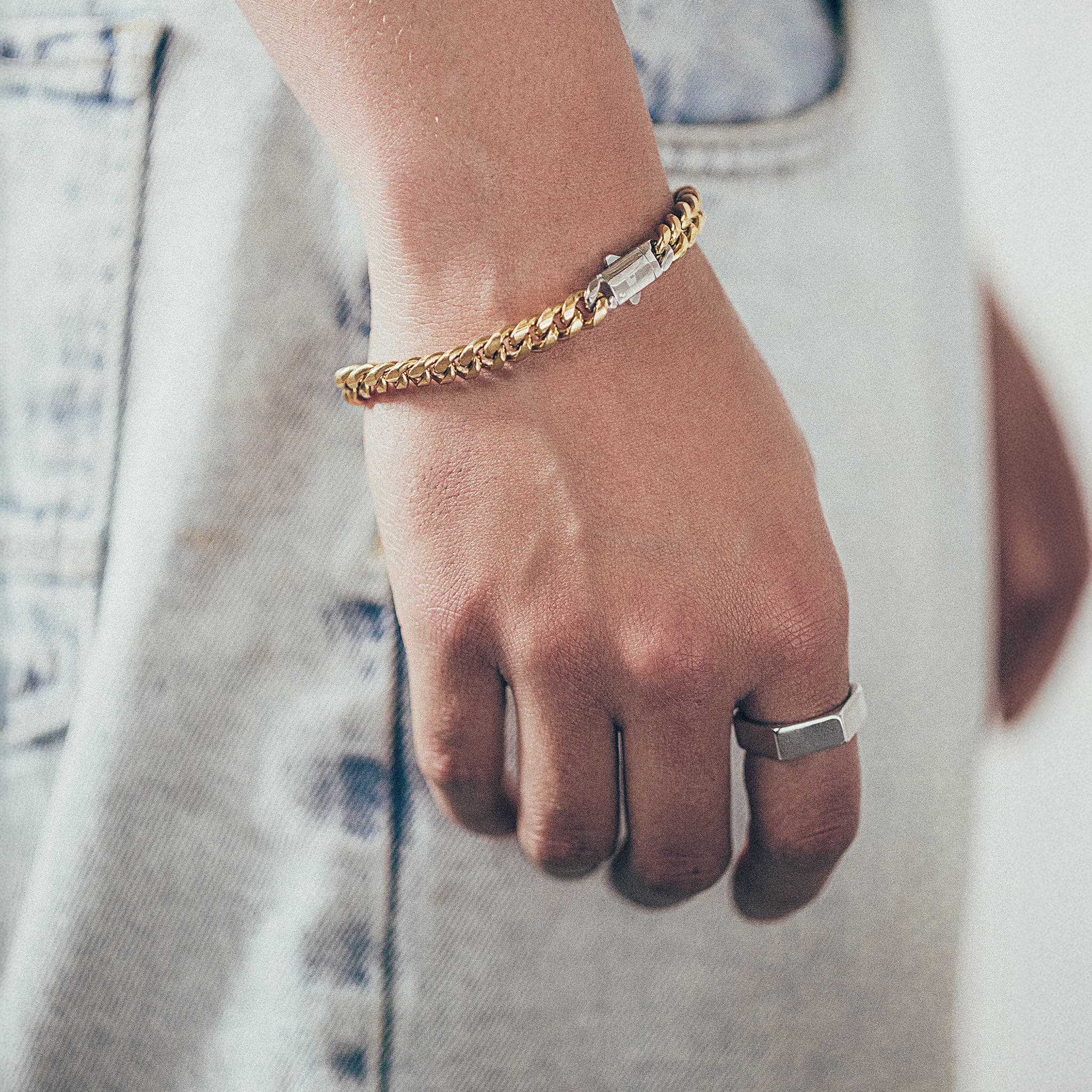 Close-up of hand and wrist featuring thick gold chain bracelet with silver clasp and silver hex style ring on textured fabric background