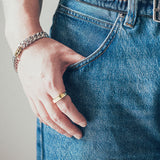 Close-up of hand and wrist featuring thick silver chain bracelet with gold clasp and gold rectangular signet-style ring on textured fabric background