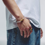 A close-up of a person's hand resting in a blue denim pocket, wearing a silver chain bracelet with a gold hex magnetic clasp and a gold ring on the finger, against a light gray background.