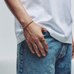 A close-up lifestyle shot of a person wearing a gold ball chain bracelet on their wrist and a gold ring on their finger, paired with a white t-shirt and blue jeans.