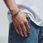 A close-up lifestyle shot of a person's hand resting on their blue jeans, showcasing a gold ball chain bracelet with a silver hexagonal block, magnetic clasp on the wrist and a matching gold ring on the finger.