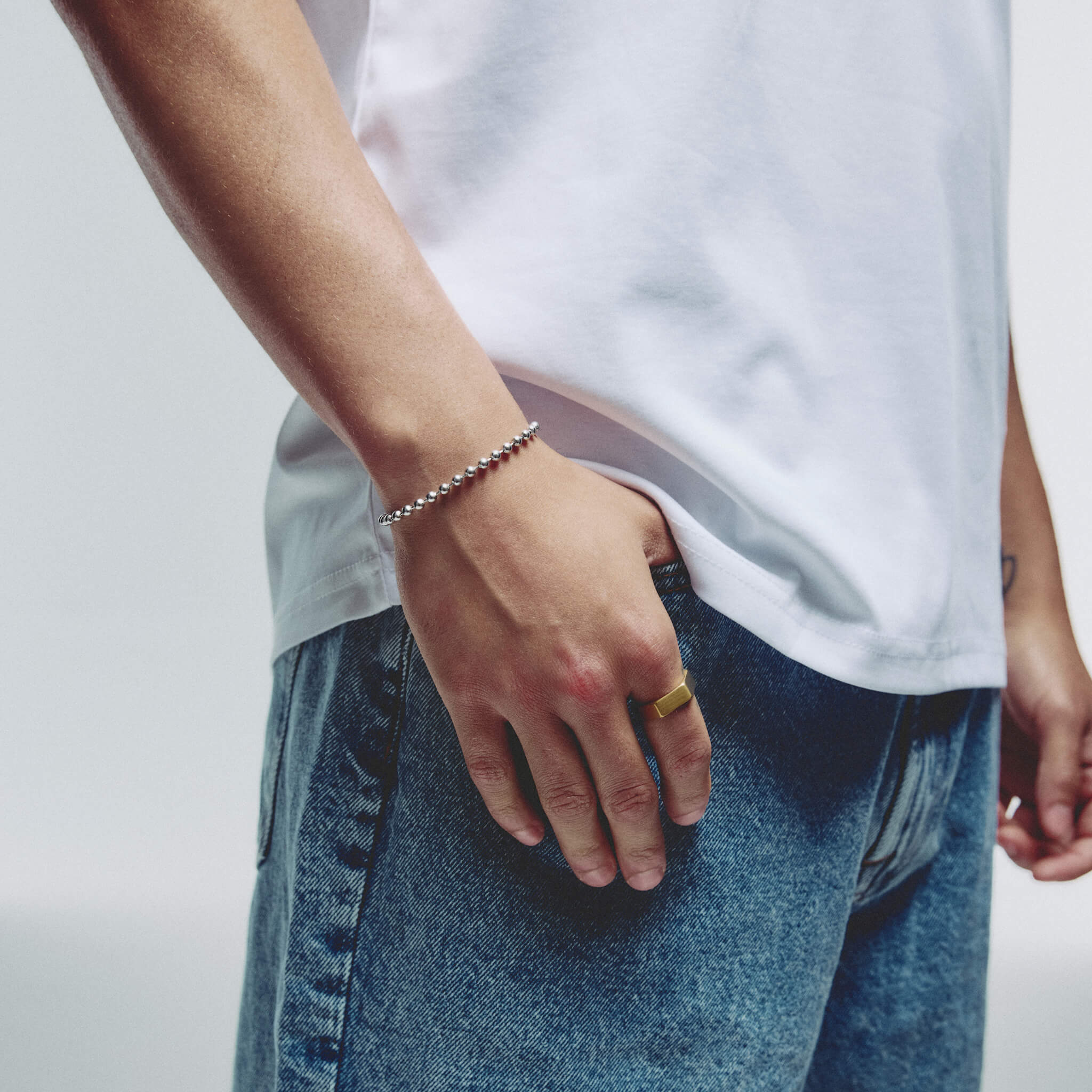 A close-up of a person wearing a delicate silver ball chain bracelet on their wrist, paired with a white t-shirt and blue jeans.