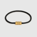 A black braided cord bracelet with a polished gold-tone triple-block hex clasp featuring an engraved 'L' logo, set against a light gray background.