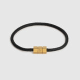 A black braided cord bracelet with a polished gold-tone triple-block hex clasp featuring an engraved 'L' logo, set against a light gray background.