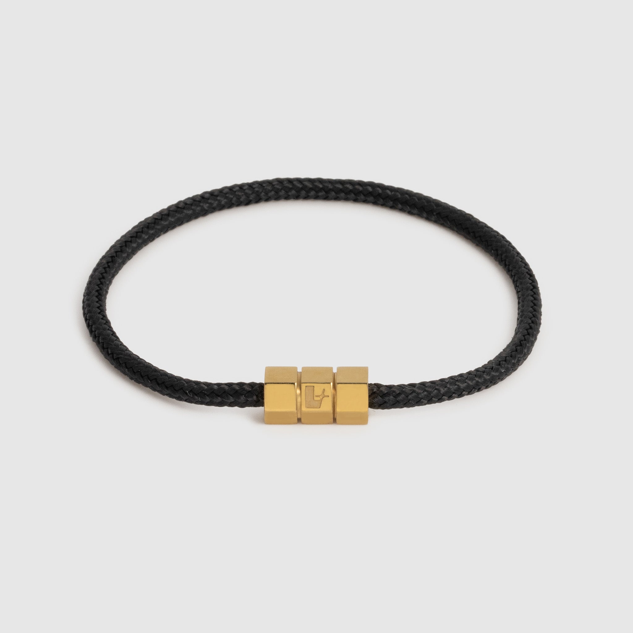 A black braided cord bracelet with a polished gold-tone triple-block hex clasp featuring an engraved 'L' logo, set against a light gray background.