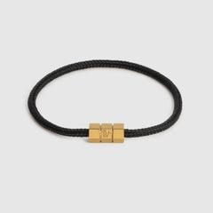 A black braided cord bracelet with a polished gold-tone triple-block hex clasp featuring an engraved 'L' logo, set against a light gray background.