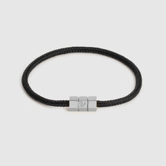 A black braided cord bracelet with a silver-toned hex block magnetic clasp featuring an engraved "L", shown on a light gray background.