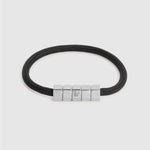 A black cord bracelet with a polished silver-toned geometric block clasp, shown on a light gray background.