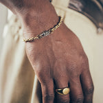 Close-up of hand and wrist featuring gold mariner-link bracelet with silver oval clasp and gold signet ring on textured fabric background”