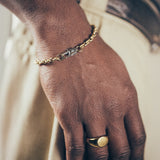 Close-up of hand and wrist featuring gold mariner-link bracelet with silver oval clasp and gold signet ring on textured fabric background”
