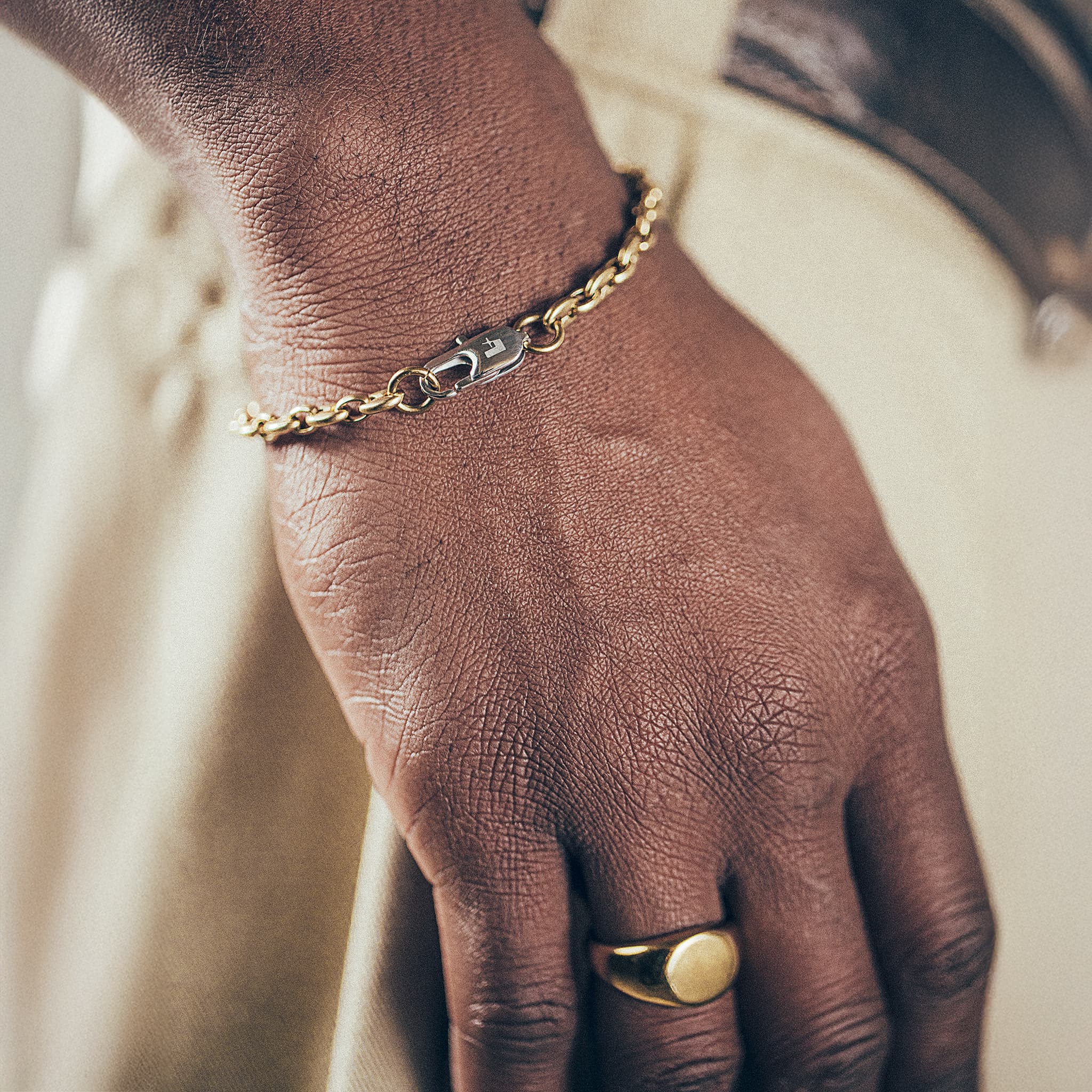 Close-up of hand and wrist featuring gold mariner-link bracelet with silver oval clasp and gold signet ring on textured fabric background”