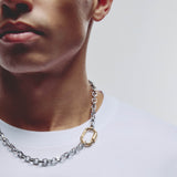 A close-up of a man wearing a silver chain necklace with a gold-toned oval clasp, paired with a white t-shirt against a light gray background.