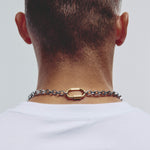 A close-up back view of a man wearing a silver-toned chain necklace with a gold-toned oval clasp, over a white t-shirt against a light gray background.