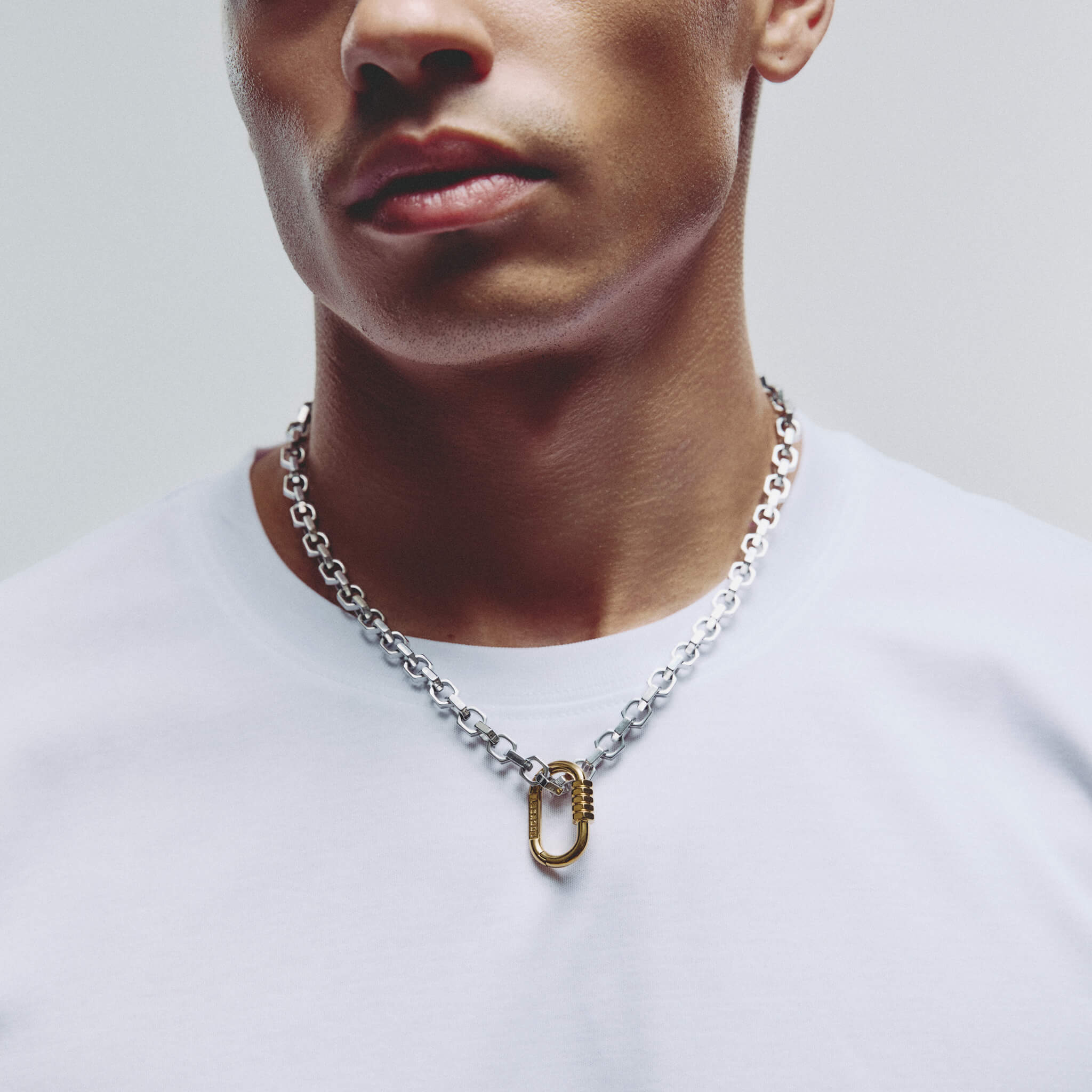 A close-up of a man wearing a silver chain necklace with a gold-toned oval lock pendant, paired with a white t-shirt against a light gray background.