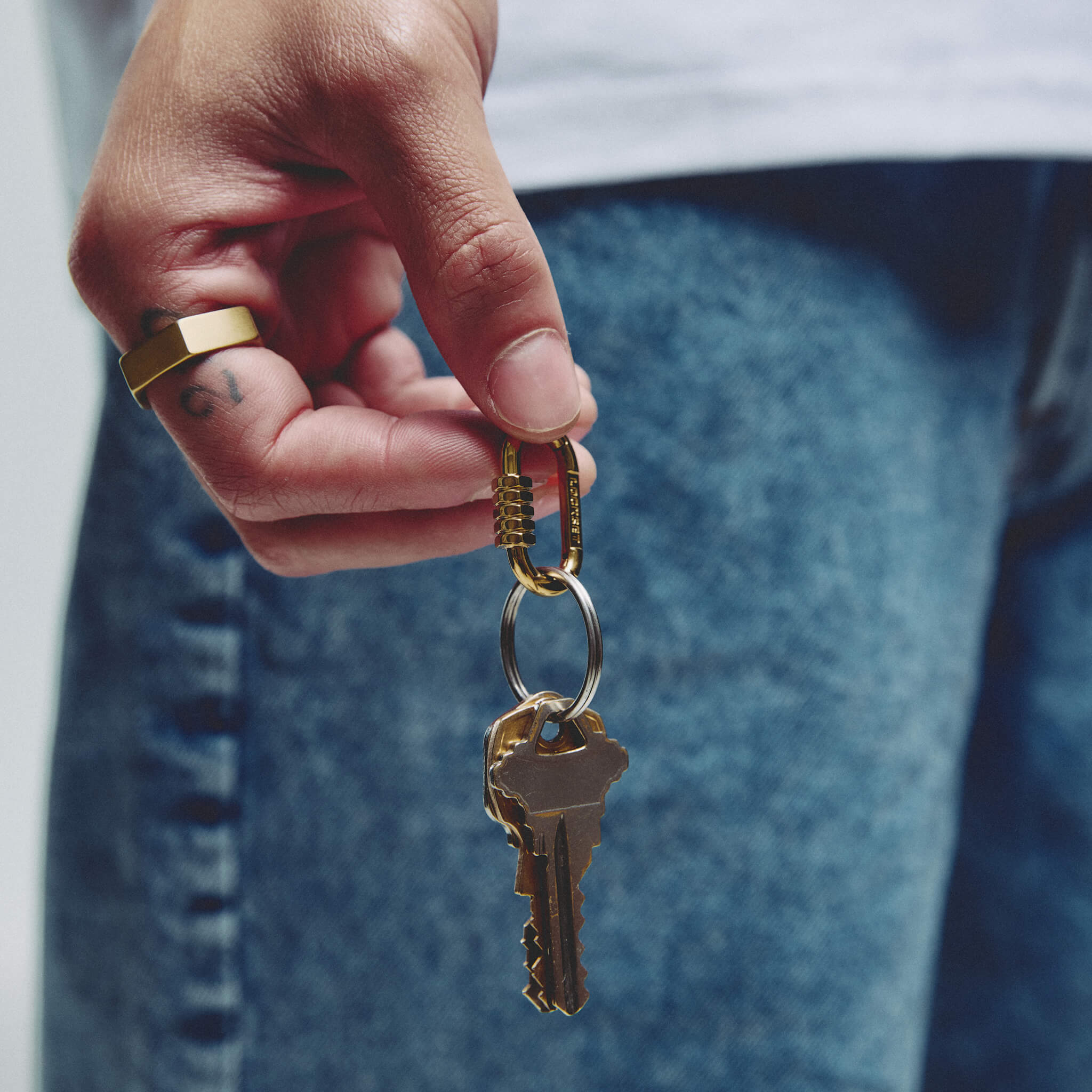 A close-up of a hand holding a brass keyring with two keys and a small combination lock, against a blurred background of blue jeans and a white shirt.
