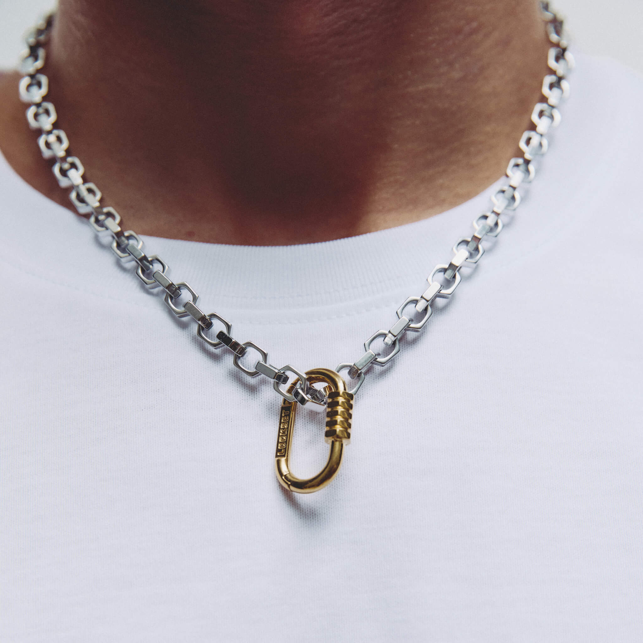 A close-up of a silver chain necklace with a gold carabiner pendant, worn on a person's neck over a white t-shirt.