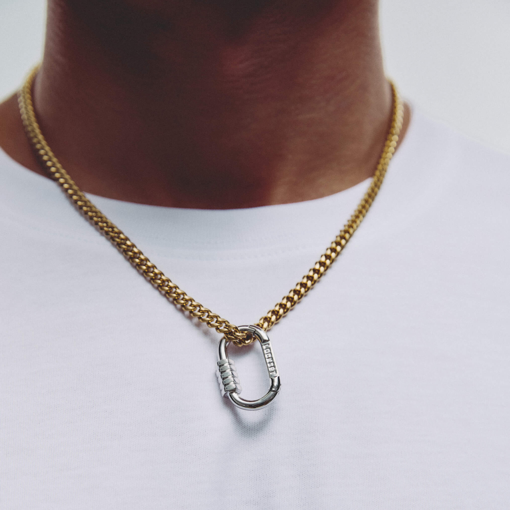 A gold chain necklace with a silver carabiner pendant, worn on a person's neck over a white t-shirt.