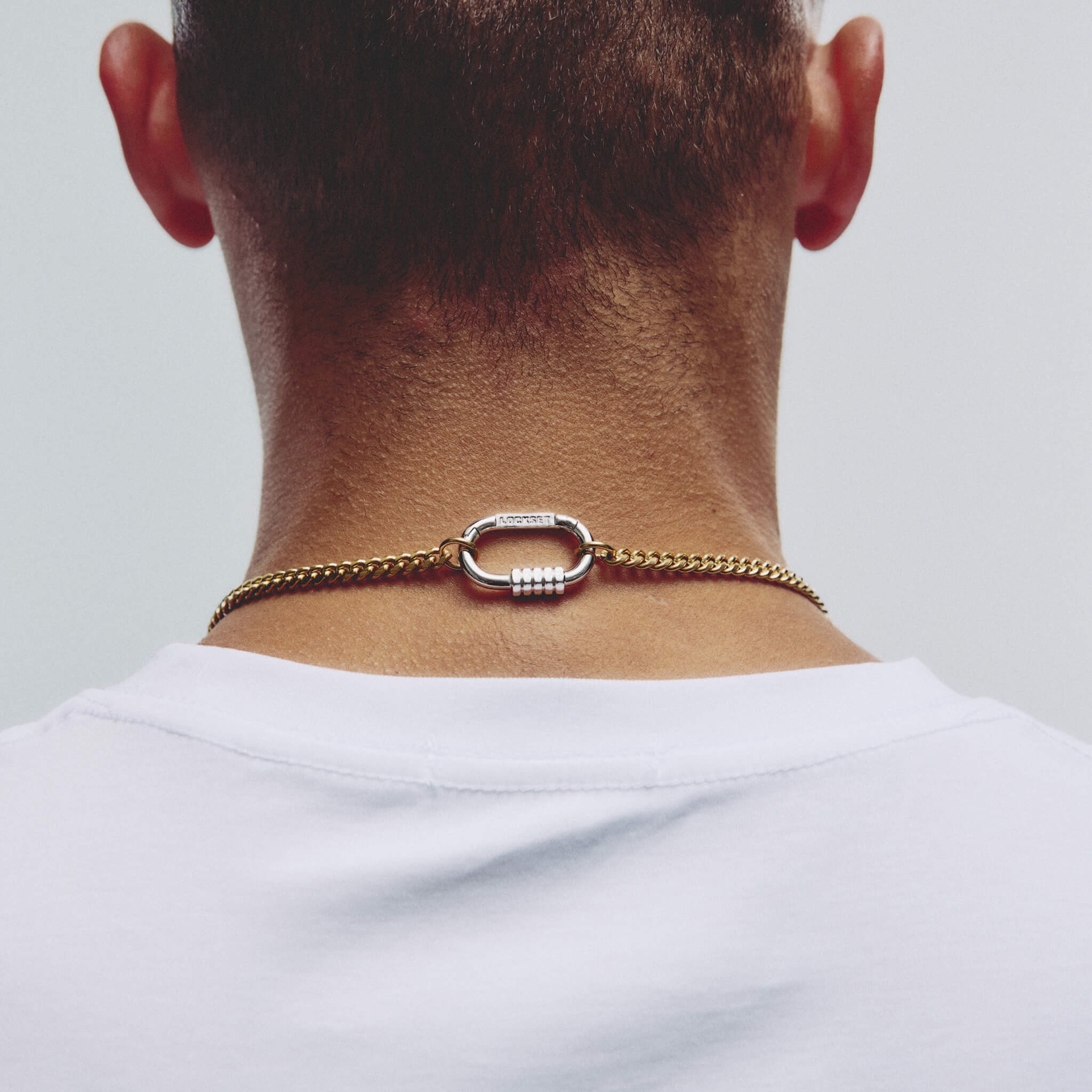 A close-up back view of a man's neck and upper shoulders, wearing a gold chain necklace with a textured silver-toned oval clasp over a white t-shirt.