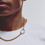 A close-up of a person wearing a gold chain necklace with a silver-toned oval link accent, paired with a white t-shirt against a light gray background.