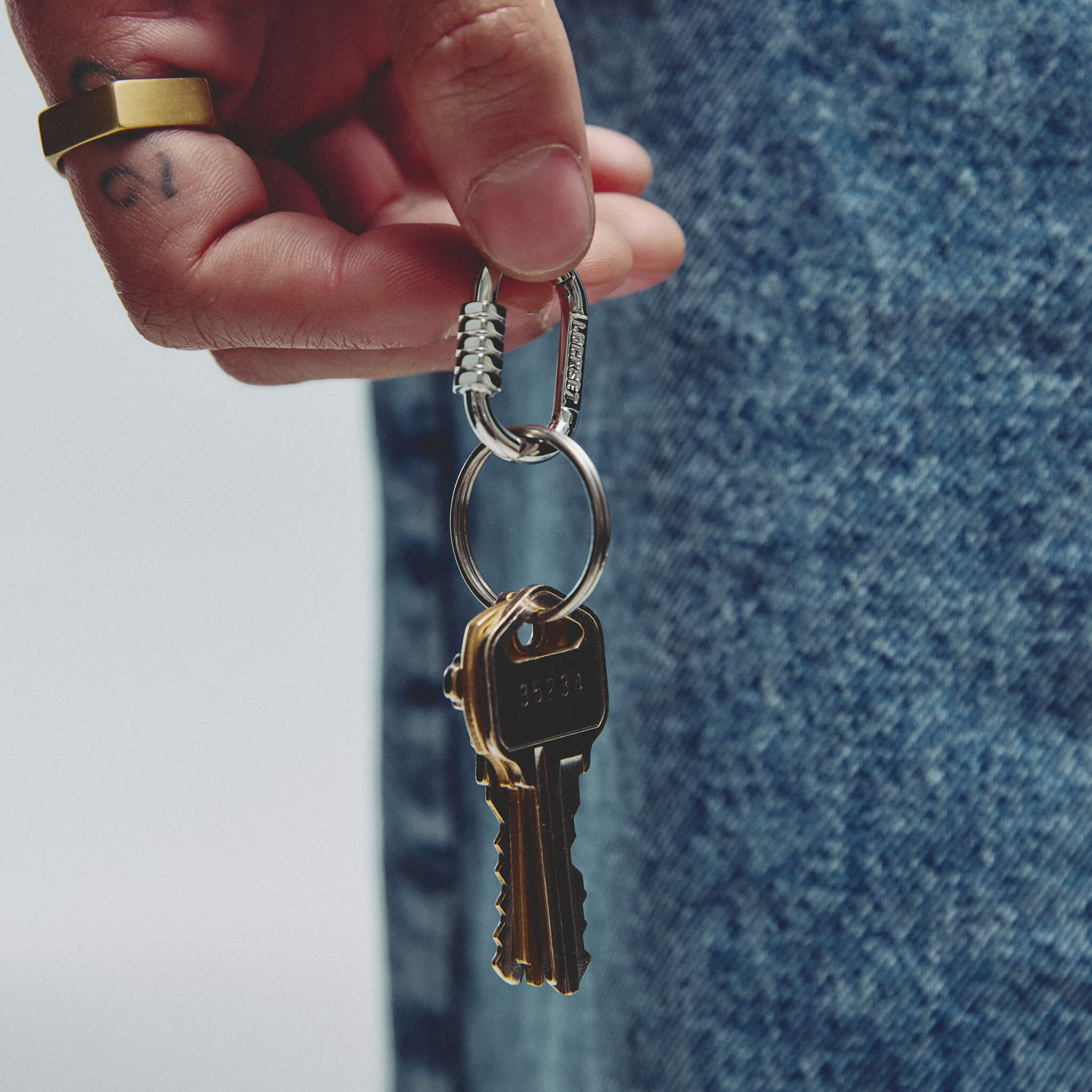 A close-up of a hand holding a silver keyring with a brass key, set against a background of blue denim jeans and a white wall.