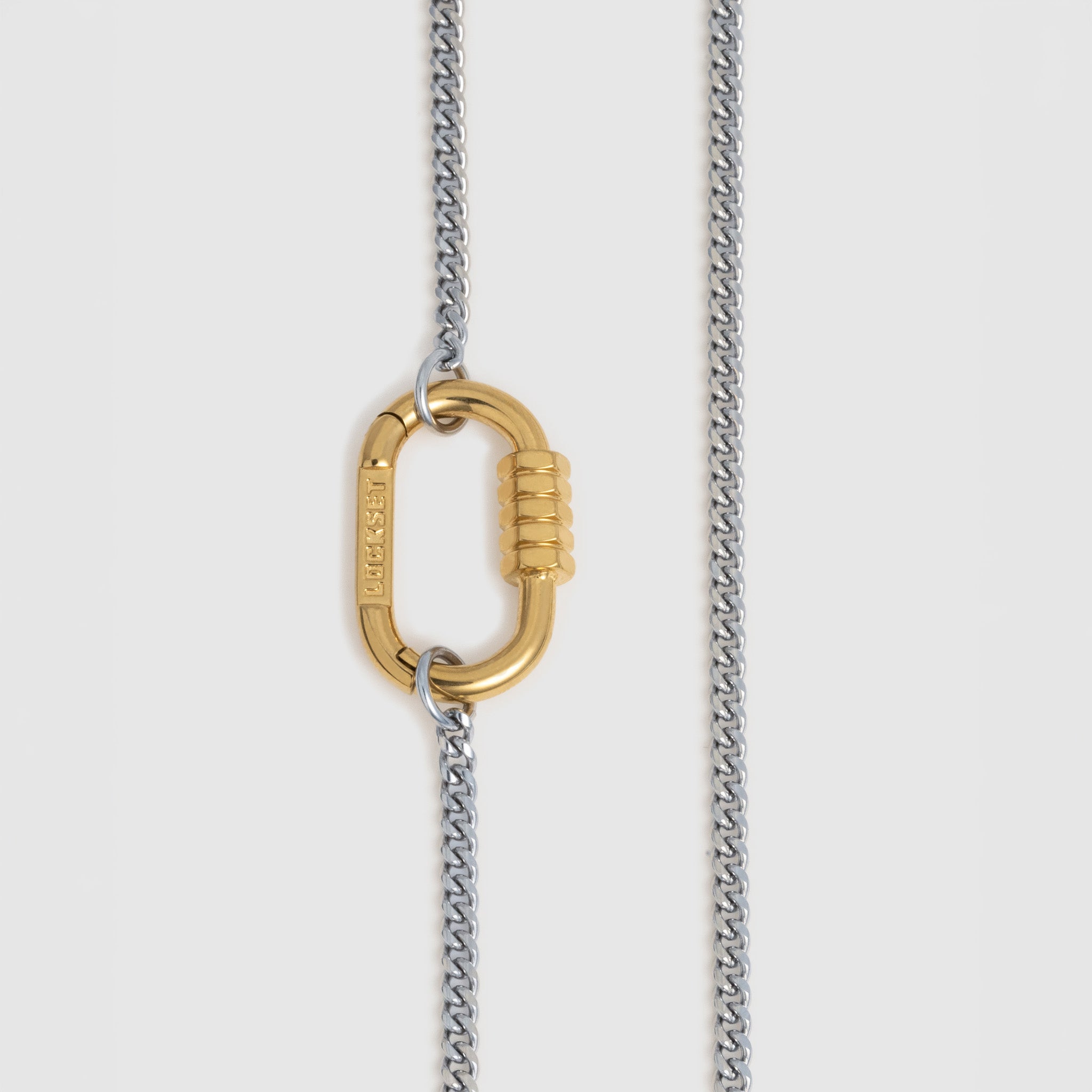A close-up of a necklace featuring a gold carabiner clasp hanging from a two-tone chain with silver and gold links, set against a light gray background.