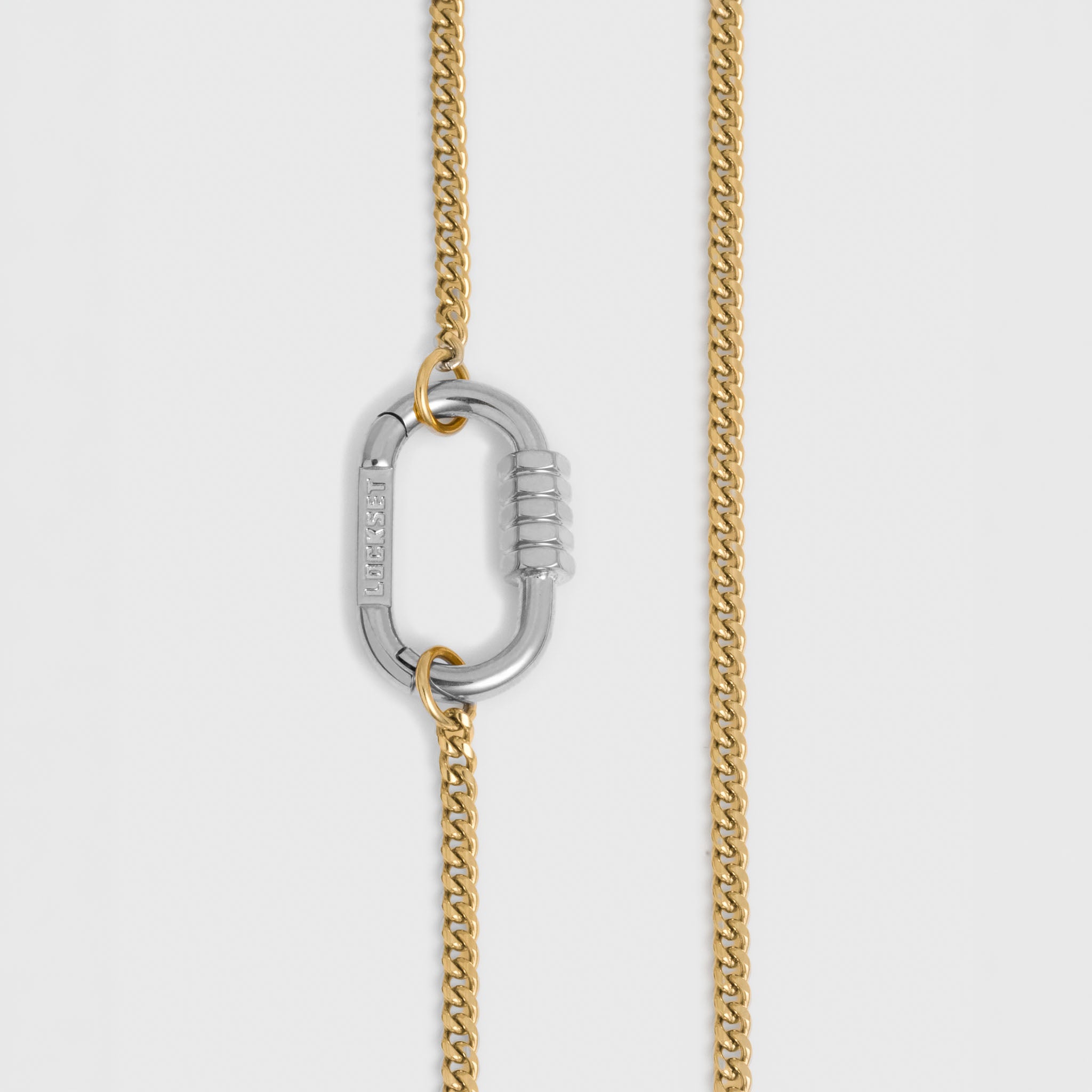A close-up of a gold-toned chain necklace with a silver-toned carabiner clasp, shown against a light gray background.
