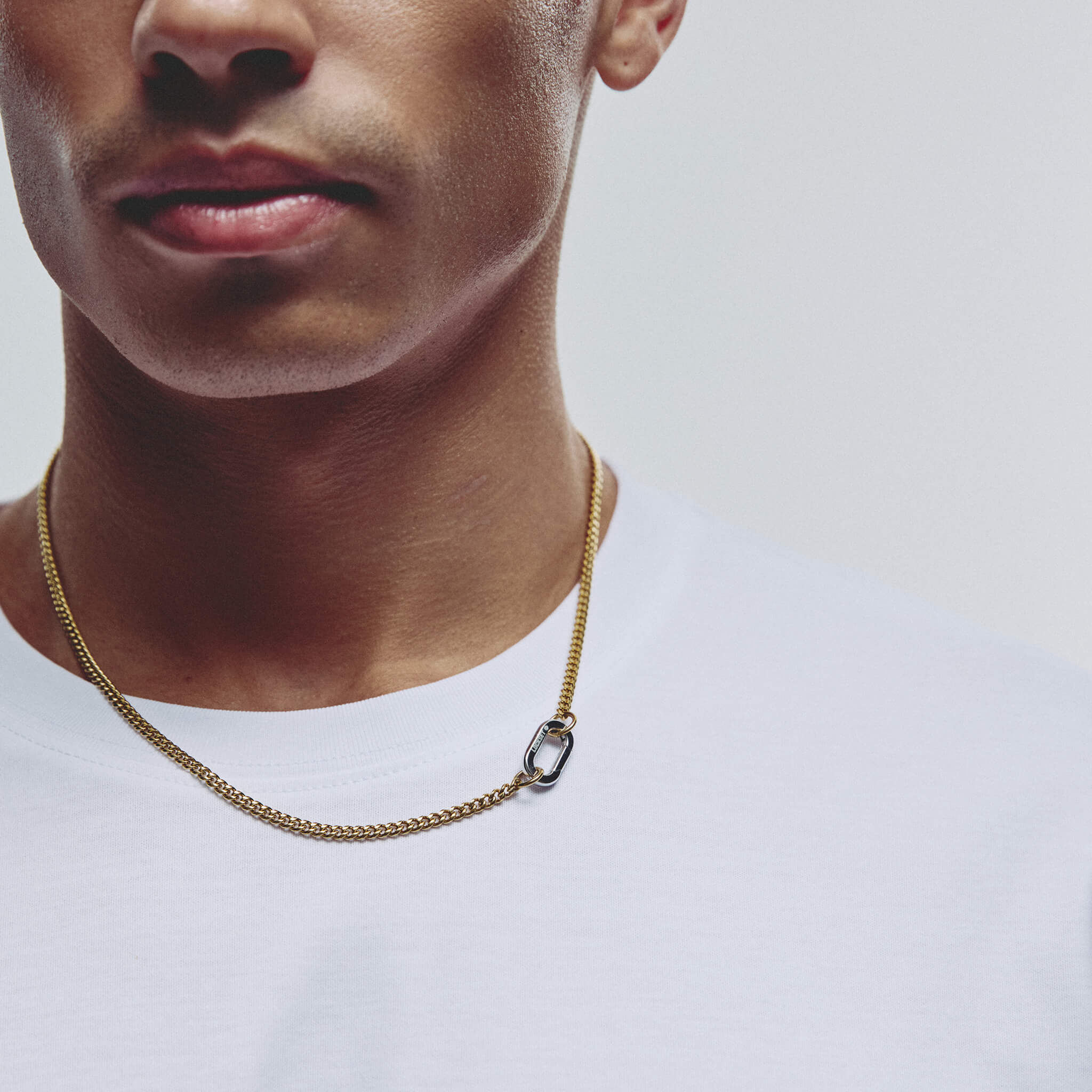 A close-up of a man wearing a gold chain necklace with a small carabiner clasp, paired with a white t-shirt against a light gray background.