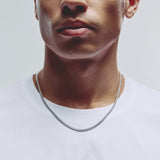 A close-up of a man wearing a silver chain necklace with a box link style, paired with a white crew-neck t-shirt against a light gray background.