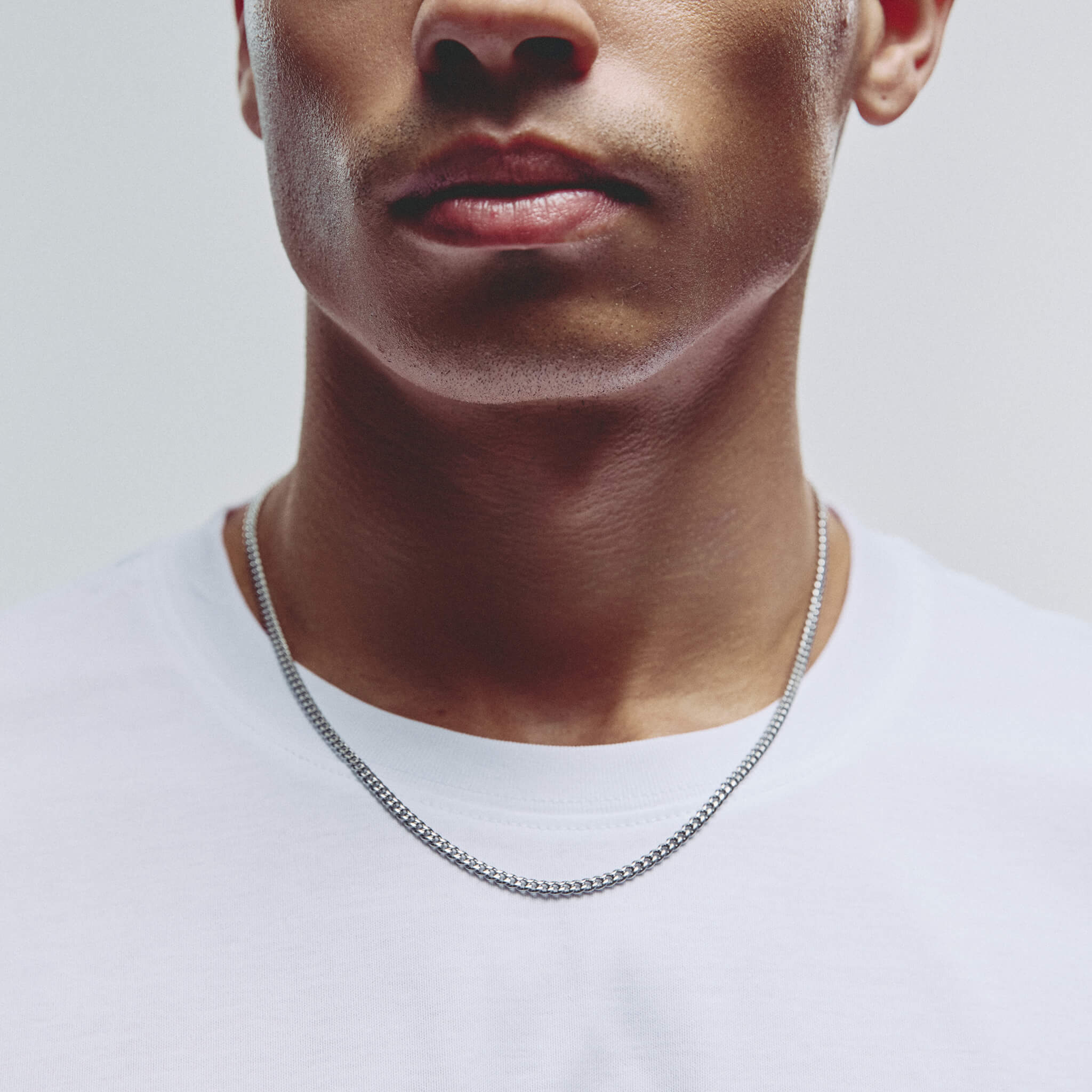 A close-up of a man wearing a silver chain necklace with a box link style, paired with a white crew-neck t-shirt against a light gray background.