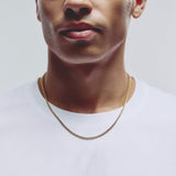 A close-up of a man wearing a delicate gold chain necklace over a white t-shirt, cropped from the nose down against a light gray background.